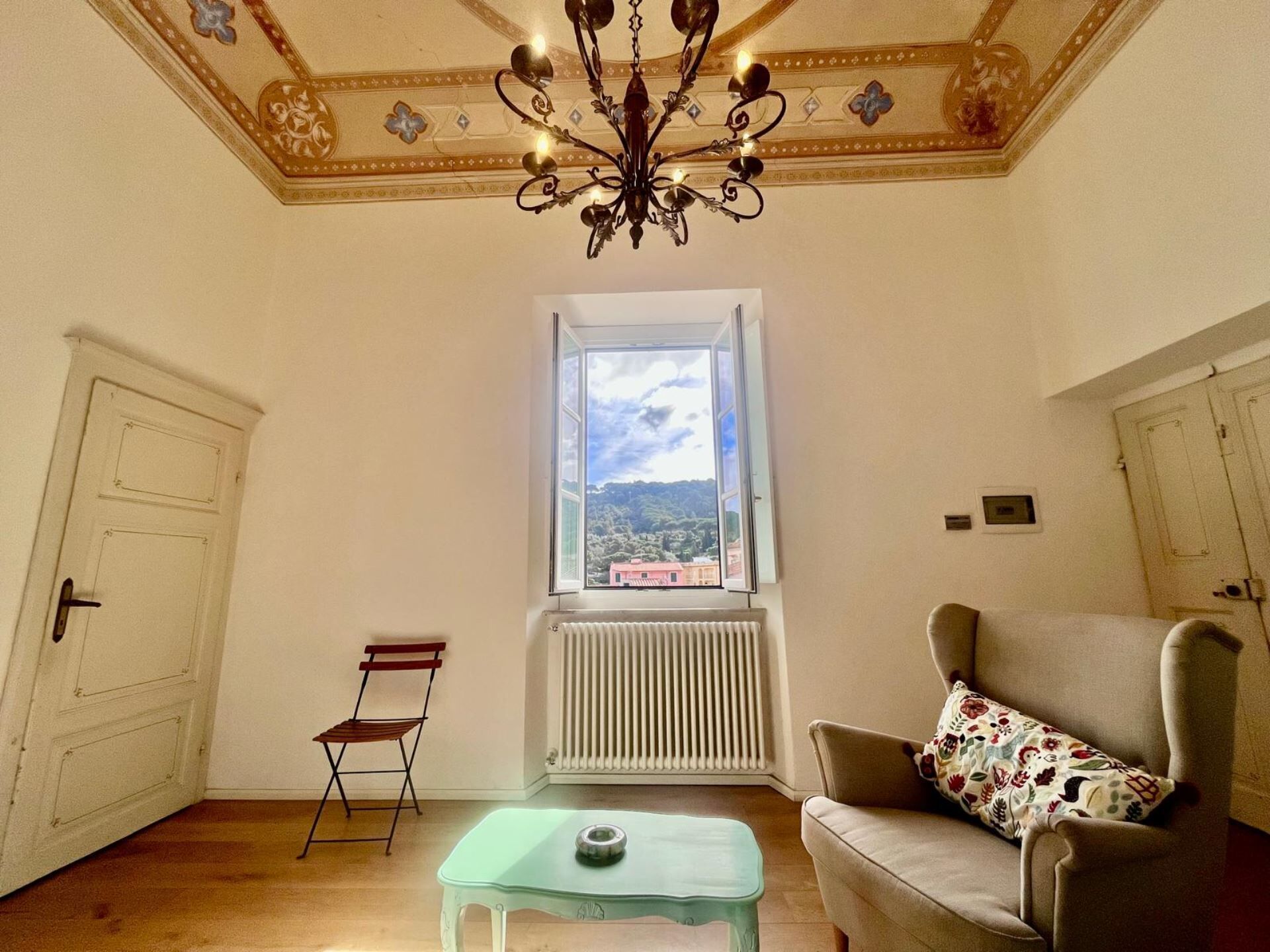 Une pièce avec une grande fenêtre, un radiateur, un lustre, un canapé et une petite table.