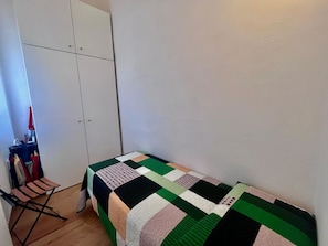 Ein kleines Zimmer mit einem Einzelbett, einer grün-weißen Steudecke, einem Holzstuhl und einem weißen Kleiderschrank.