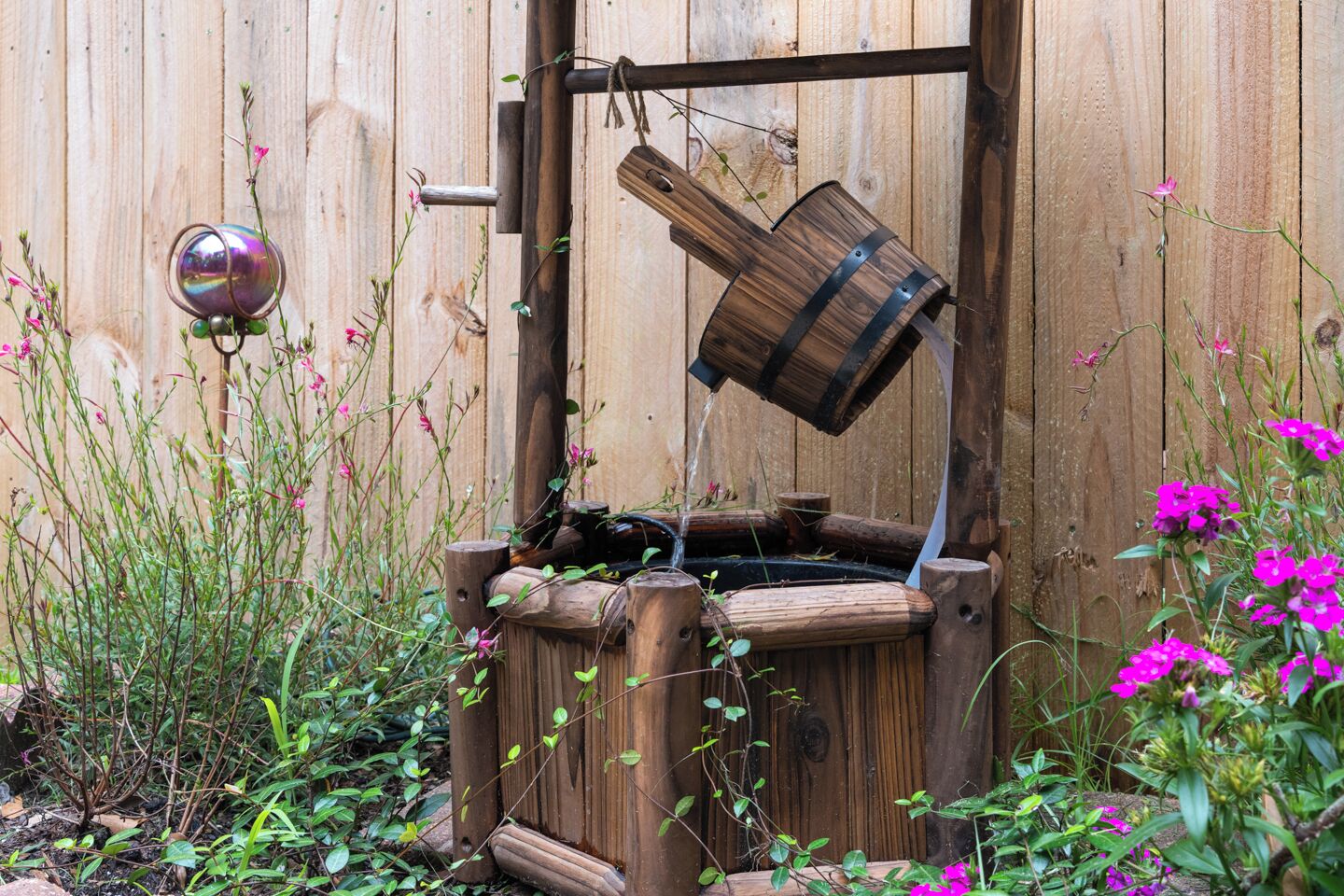 Una estructura rústica de madera con una fuente, rodeada de follaje y flores.