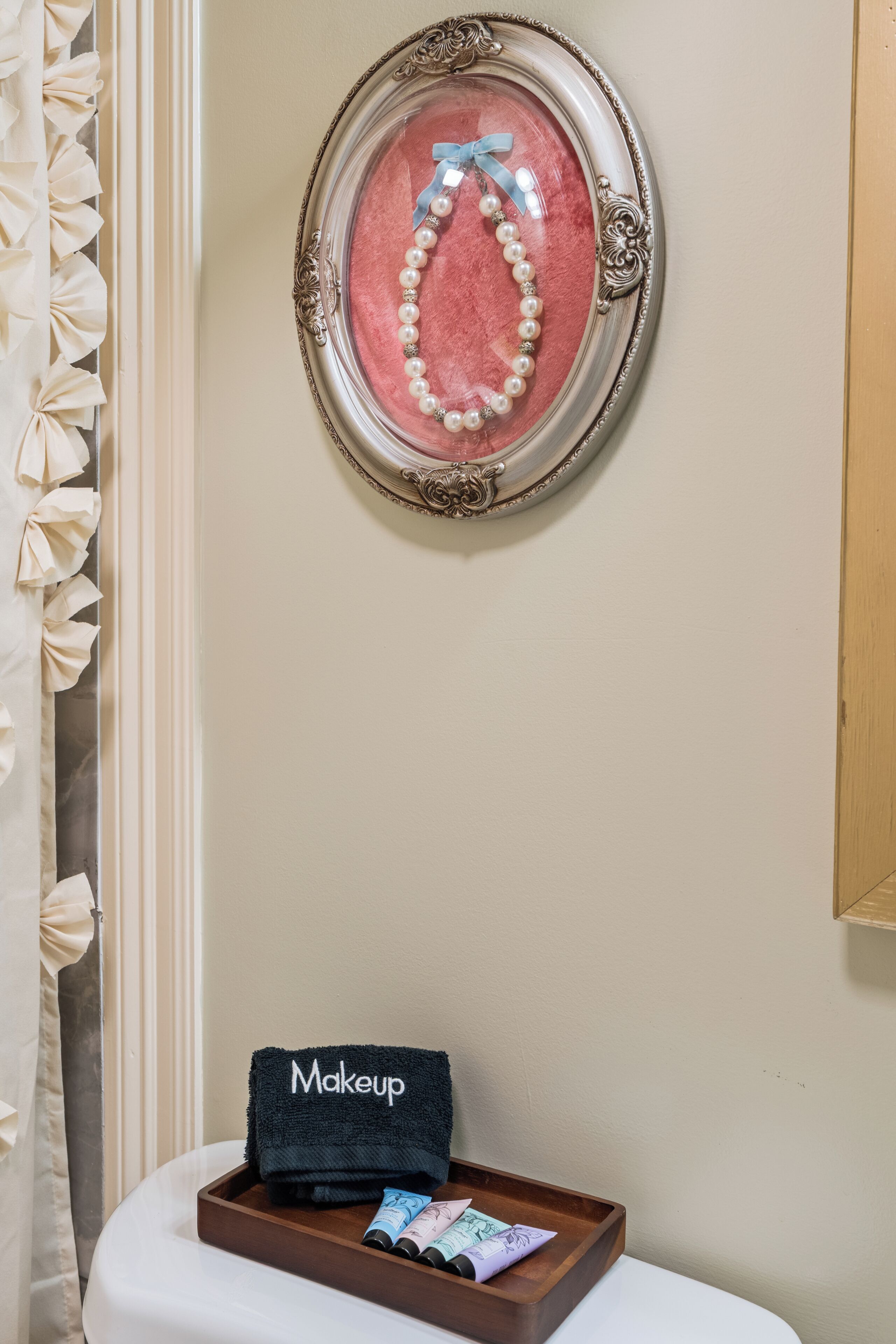 Un collar de perlas en un marco en la pared, un neceser con la etiqueta 'Makeup' sobre una mesita y una bandeja de madera con productos de cosmética.