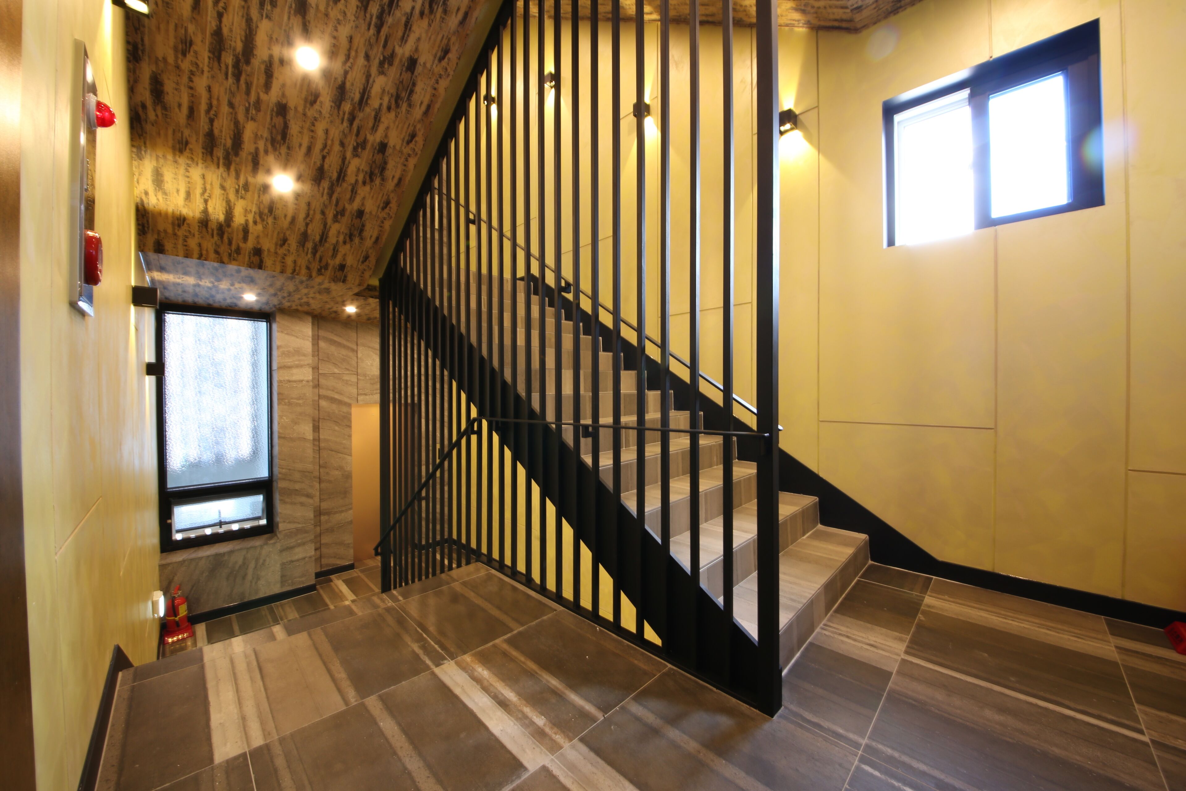 Una escalera con barandilla negra, una ventana a la izquierda y un muro con diseño texturizado.