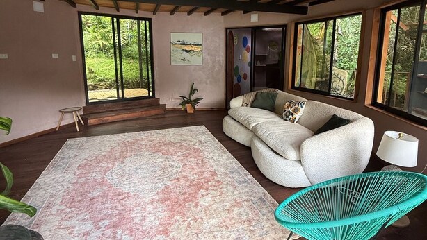 A spacious living area with a large rug, a sofa, and a chair. There are large windows providing a view of greenery outside. - Beautiful 2 Story Dome Perfect for Your Getaway (Rivas)
