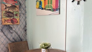 A small, enclosed patio area with a round wooden table, two wicker chairs, a wall-mounted clock, and a painting of colorful beach huts. - Doña Lucía (Guánica)