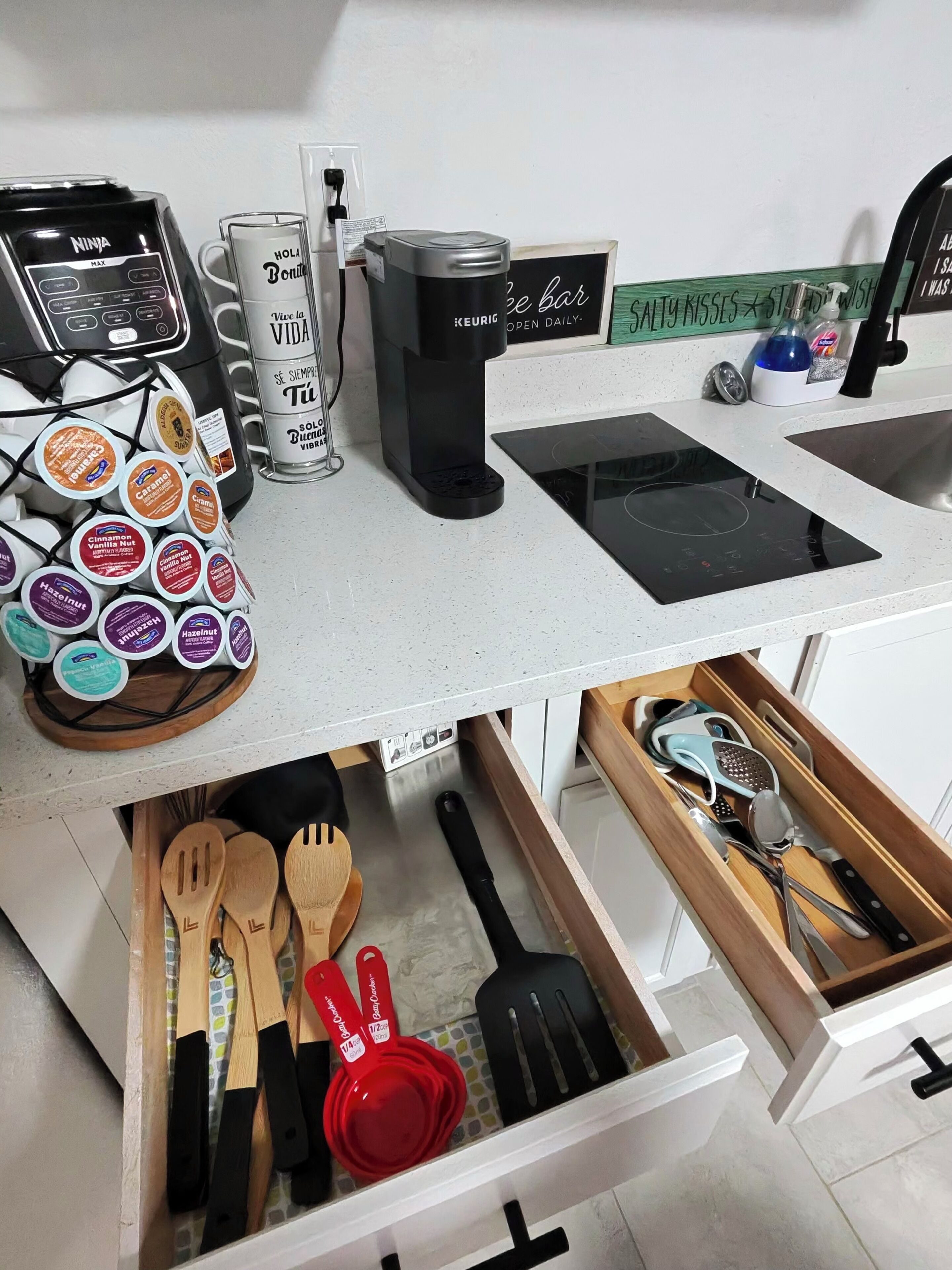 Una encimera de cocina con una cafetera, una taza, un hervidor y una máquina de café. También se ven utensilios y un especiero.