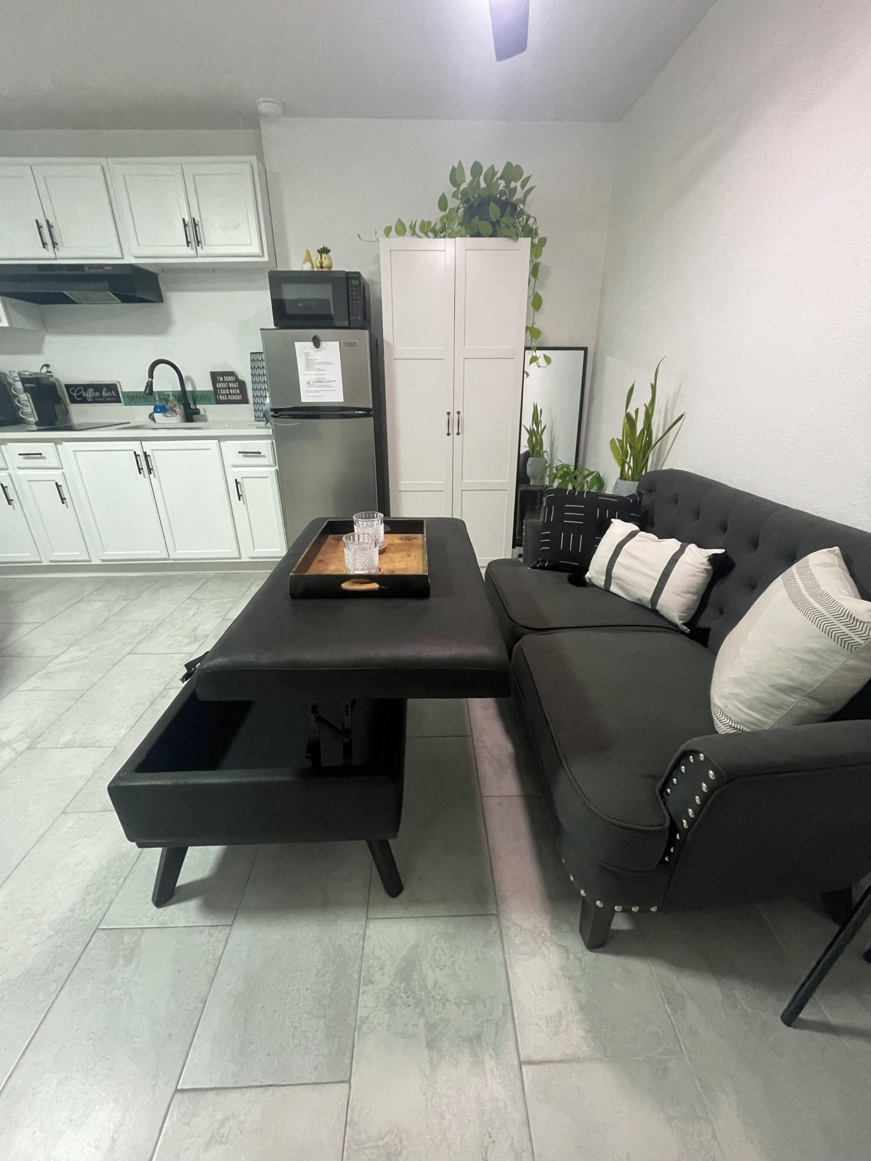 Una sala de estar moderna con un sofá negro, una mesa de centro, un mueble aparador y una cocina al fondo.