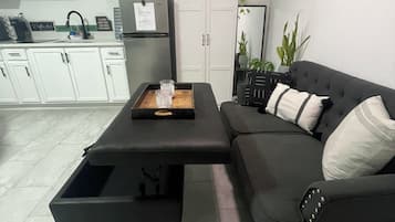 Una sala de estar moderna con un sofá negro, una mesa de centro, un mueble aparador y una cocina al fondo.