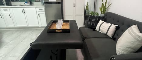 A modern living room with a black sofa, a coffee table, a cabinet, and a kitchen area in the background.