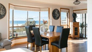 Een eethoek met een houten tafel en stoelen, grote ramen met uitzicht op zee, en een decor kast.