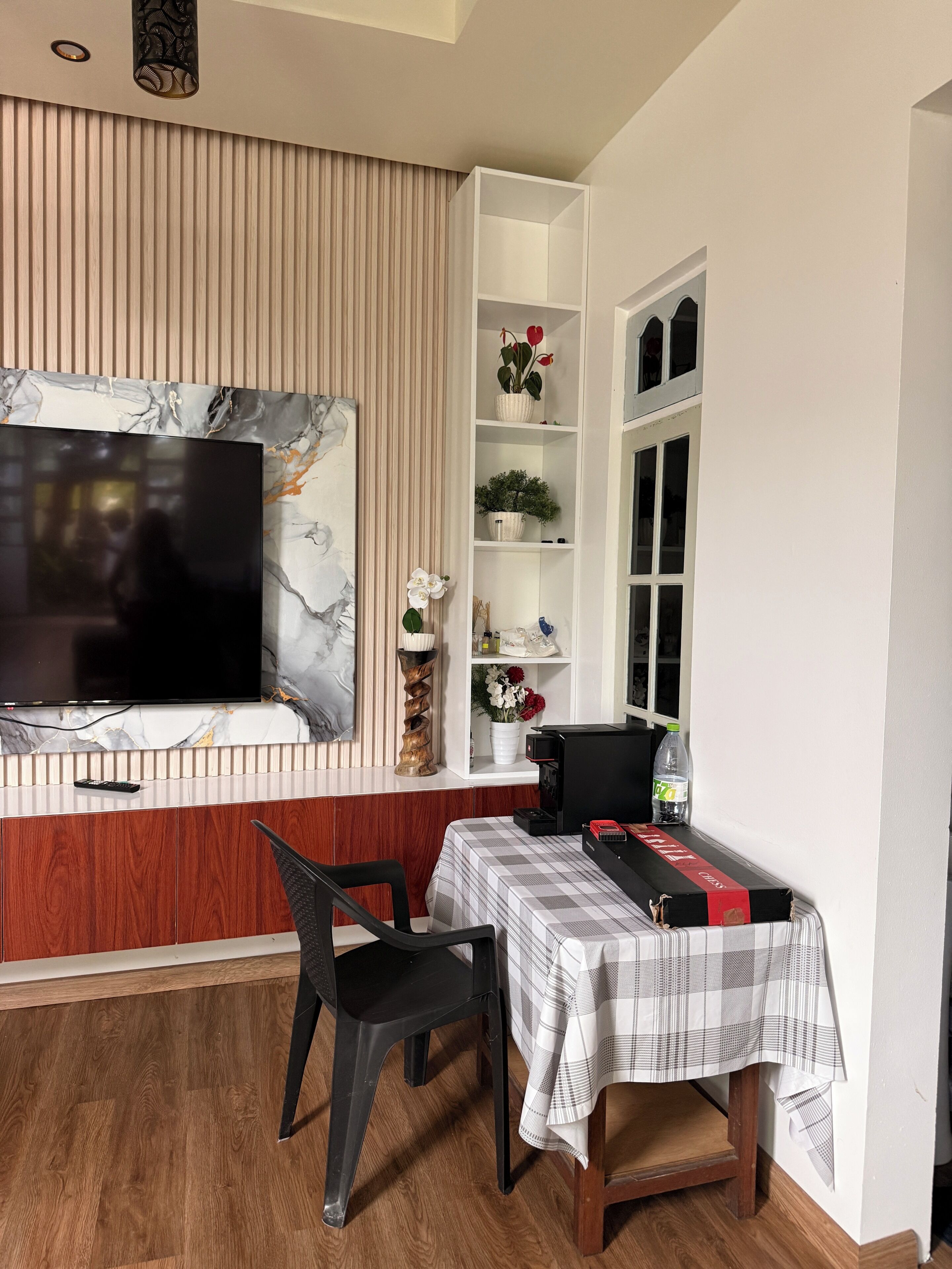 A modern living room with a TV, wooden paneling, a white shelf with decorative items, a dining area with a table and chairs, and a window with blinds.