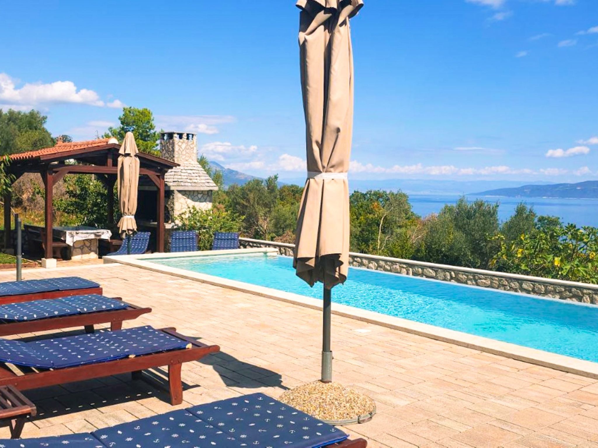 Zona de piscina con sillones de descanso y una sombrilla, un edificio de piedra y vistas al mar.