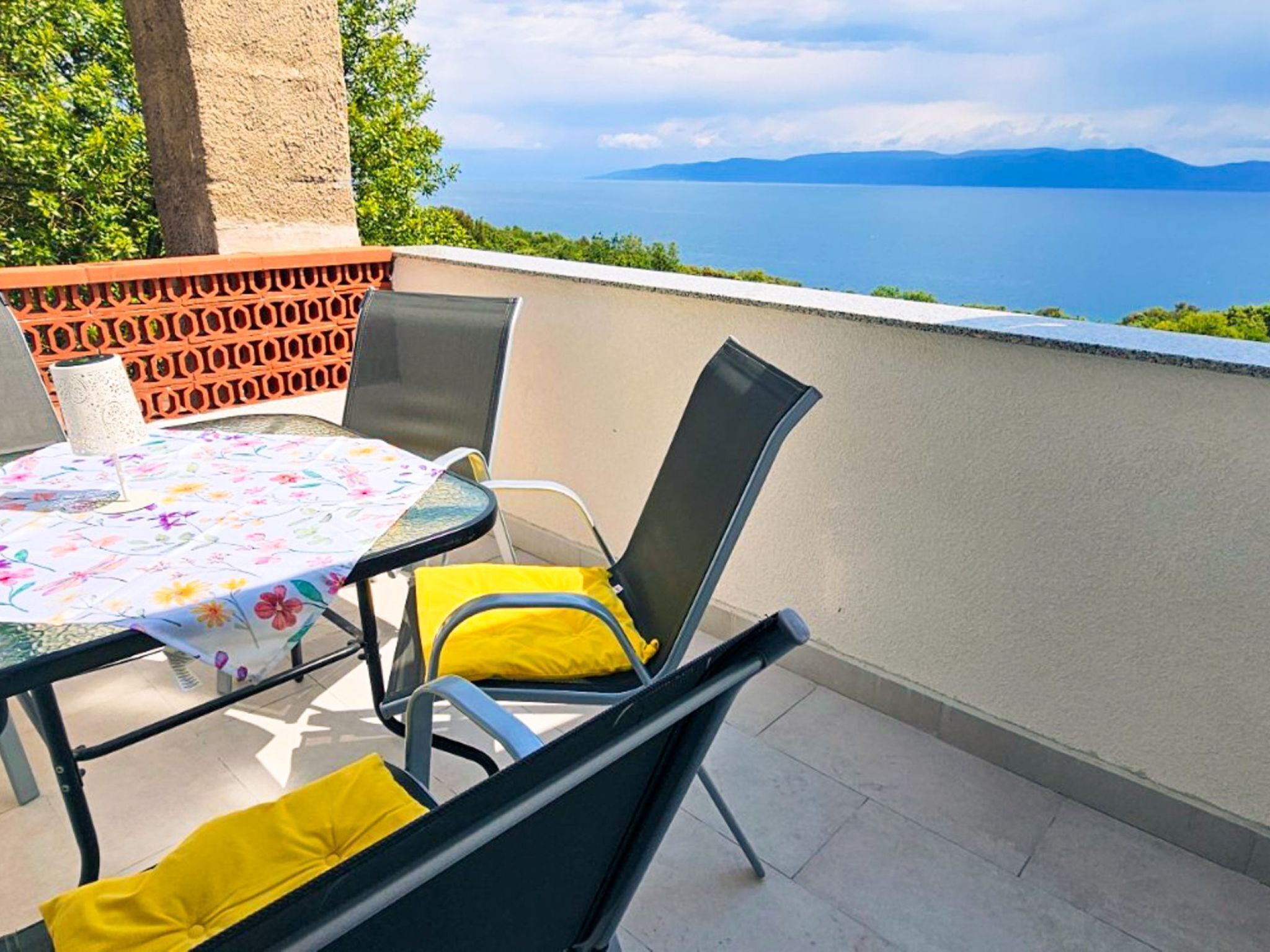A balcony with a table and chairs, a view of the sea, and a distant mountain range.