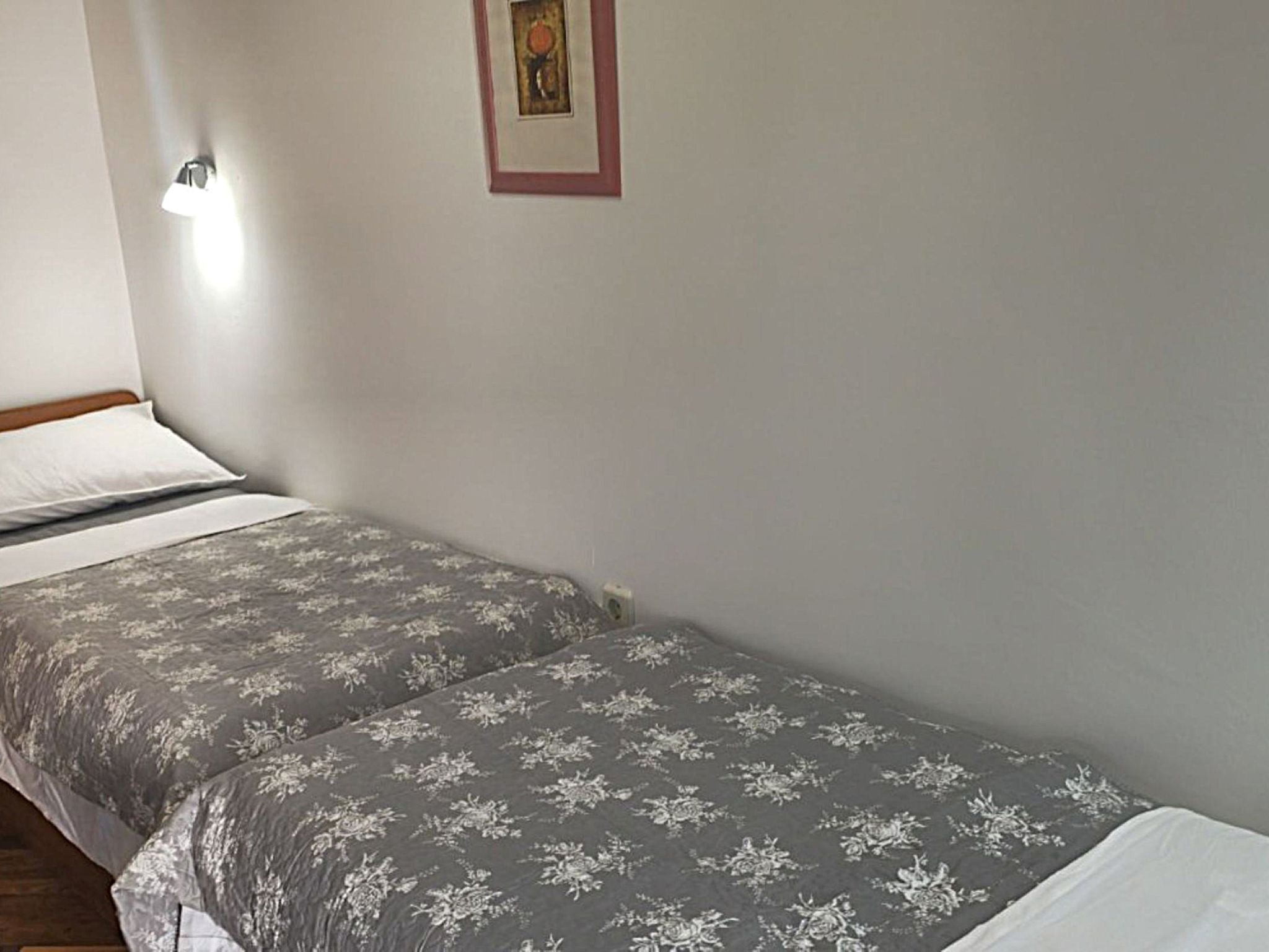 En enkeltseng med et blomstret sengetæppe, en hvid pude og en vægmonteret lampe.