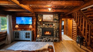 L’intérieur d’une cabane en rondins, avec une cheminée en pierre, un plancher en bois, un canapé en cuir, un téléviseur et un mur en pierre orné d’un tableau.
