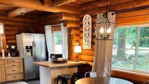 L’intérieur d’une cabane en rondins, avec une table à manger en bois, des chaises, un réfrigérateur et une fenêtre donnant sur un paysage verdoyant.