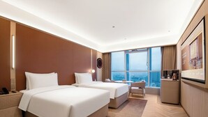 A hotel room with two beds, a desk, a chair, a small table, and a large window offering a city view.
