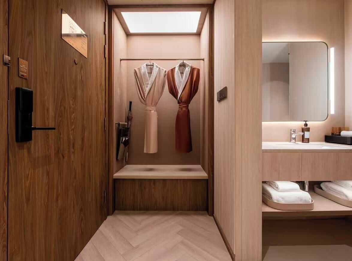 A compact, modern bathroom with wooden walls, a built-in bench, a mirror, and two hanging robes.