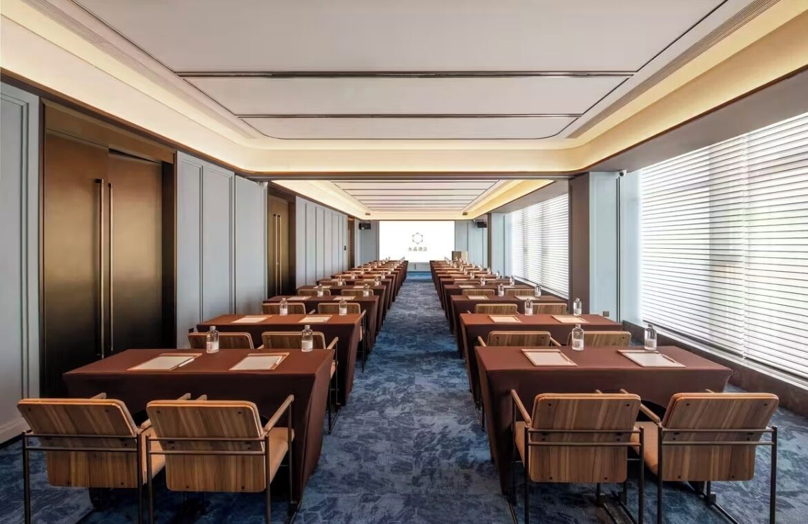 A conference room with rows of tables and chairs, a projector screen at the front, and a carpeted floor.