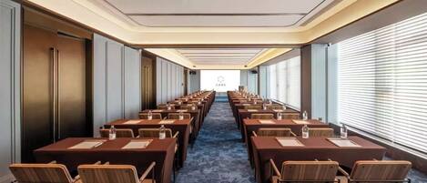 A conference room with rows of tables and chairs, a projector screen at the front, and a carpeted floor.