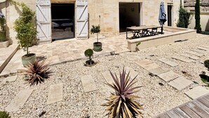 Un edifici de pedra amb diverses portes i finestres, una zona de pati amb seients i plantes en testos.