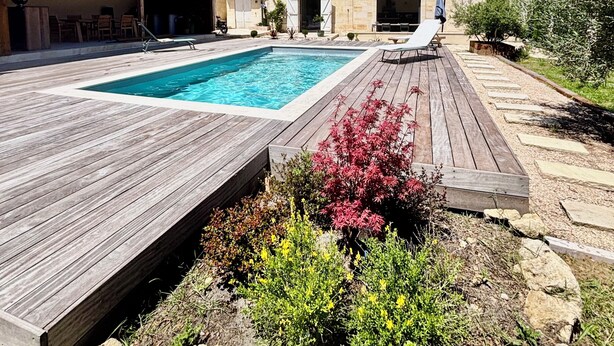 Una piscina amb una terrassa de fusta, un edifici amb teulada d'àmples rajoles i un jardí amb pedres i plantes.