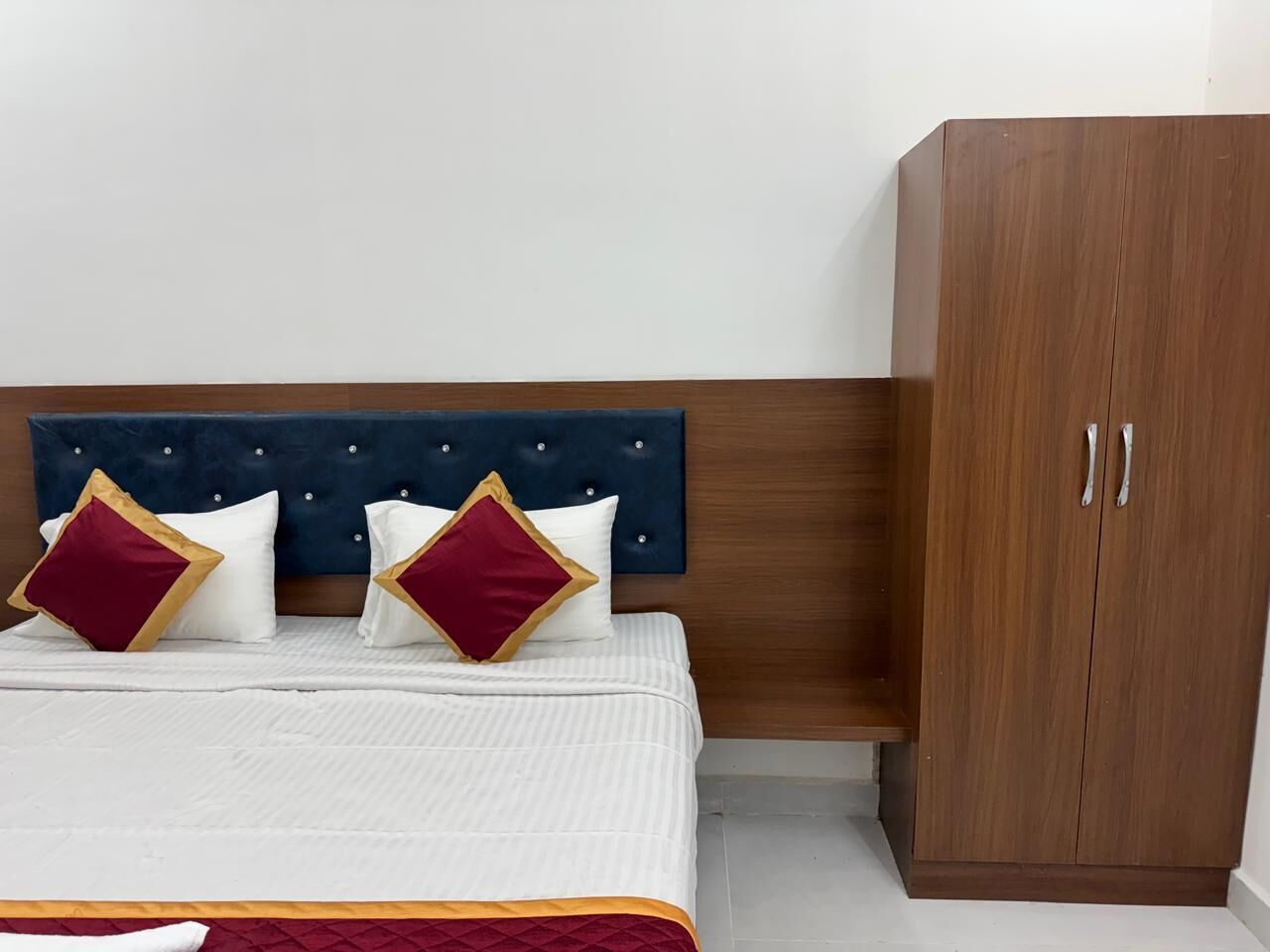 Una cama con cabecera capitonada, dos almohadas con detalles dorados y un armario de madera.