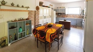 A dining area with a table set for four, surrounded by chairs. There is a kitchen area in the background with cabinets and a window. - Nature Villa · Pool · Large Garden · Cap Bon (Grombalia)