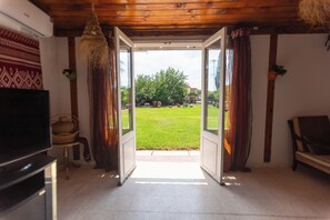An open door leading to a grassy yard with a tree, a chair, and a television in the room. - Nature Villa · Pool · Large Garden · Cap Bon (Grombalia)