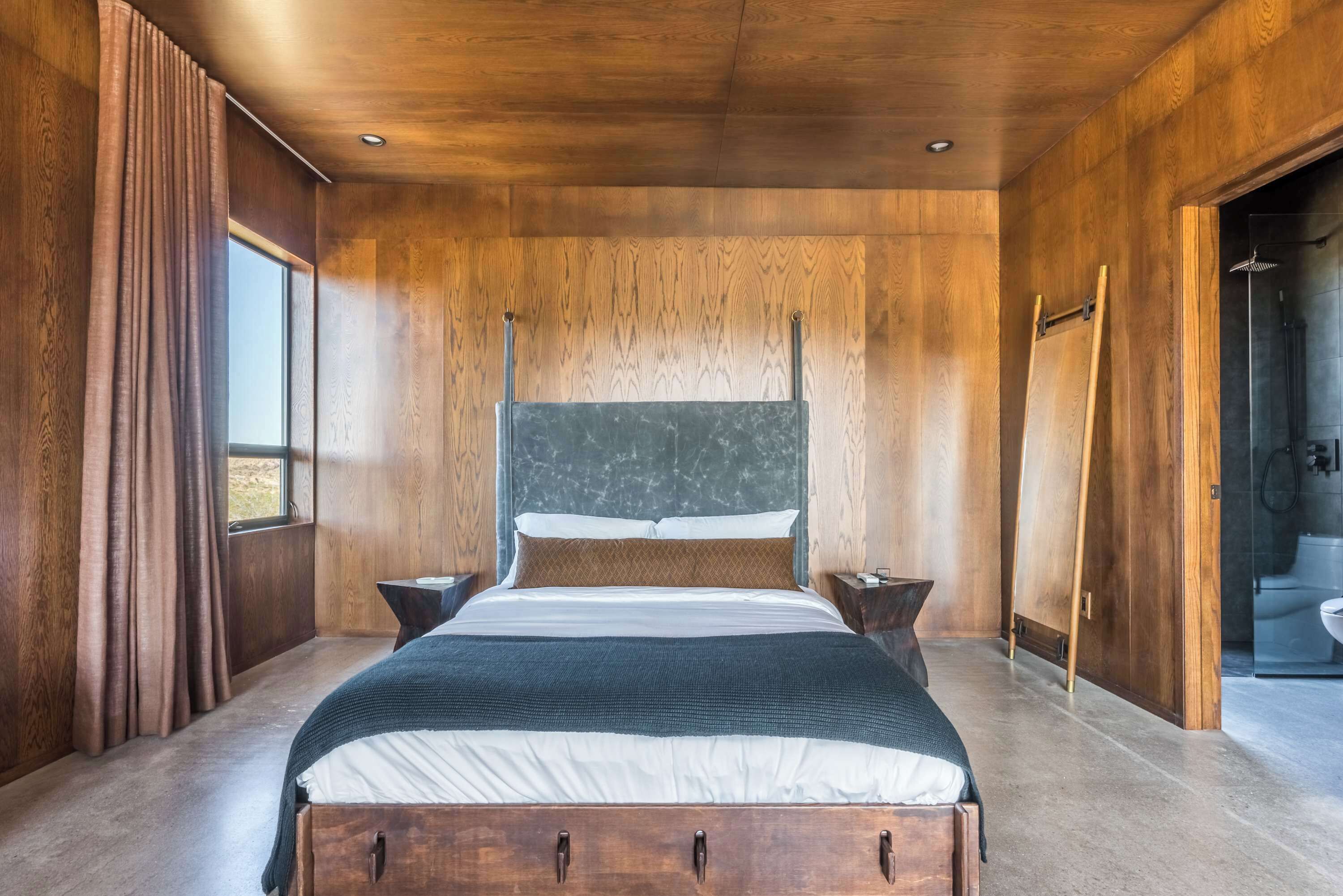 A modern bedroom with a wooden ceiling, a large bed with a headboard, two bedside tables, and a window with curtains.