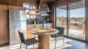 Une cuisine moderne dotée d’un réfrigérateur en acier inoxydable, de meubles en bois, d’une table ronde en bois avec des chaises, et d’une grande fenêtre offrant une vue sur le paysage désertique.