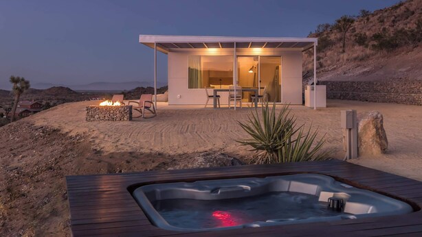 Un espace de vie extérieur moderne comprenant un jacuzzi, un foyer et une structure fermée par des parois de verre, avec une table et des chaises pour les repas.