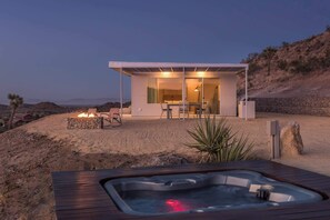 Ein moderner Außenbereich mit Whirlpool, Feuerstelle und einer gläsernen Struktur mit Esstisch und Stühlen.