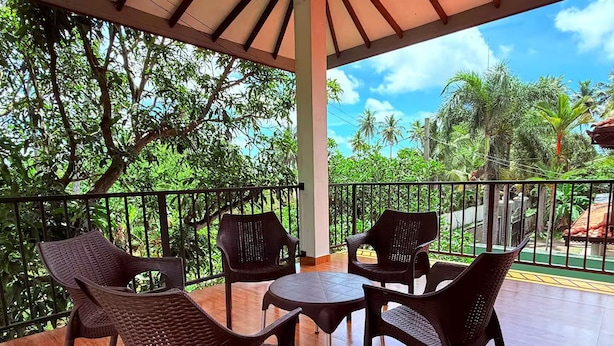 A covered outdoor seating area with wicker chairs and a table, surrounded by a metal railing and lush greenery.