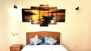 A bedroom with a bed, a nightstand, a wall-mounted painting of a sunset over water, and a wall-mounted light fixture.