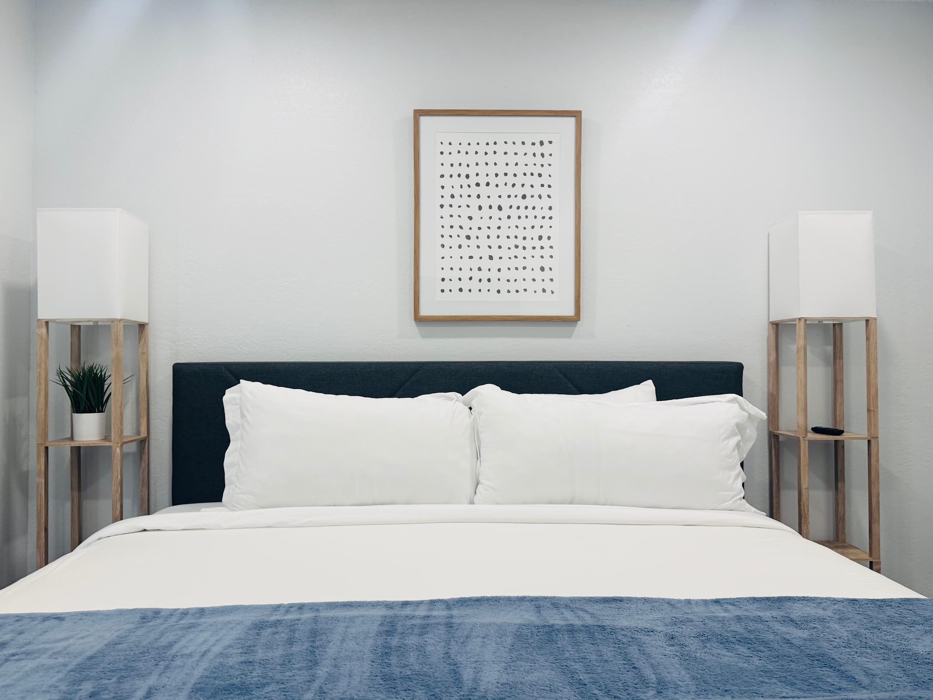 Un dormitorio ordenado con cabecera oscura, ropa de cama blanca y dos mesitas de noche con lámparas y una planta en maceta.