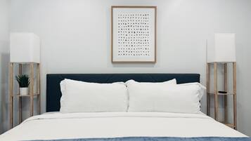 Un dormitorio ordenado con cabecera oscura, ropa de cama blanca y dos mesitas de noche con lámparas y una planta en maceta.