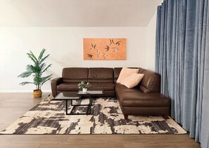 Een bruine leren bank, een bijzettafeltje, een kamerplant, een kunstwerk aan de muur en een kamerbreed tapijt in een woonkamer.