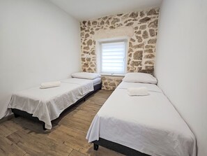 Quarto com duas camas de solteiro, parede de pedra, janela com persianas e piso de madeira.