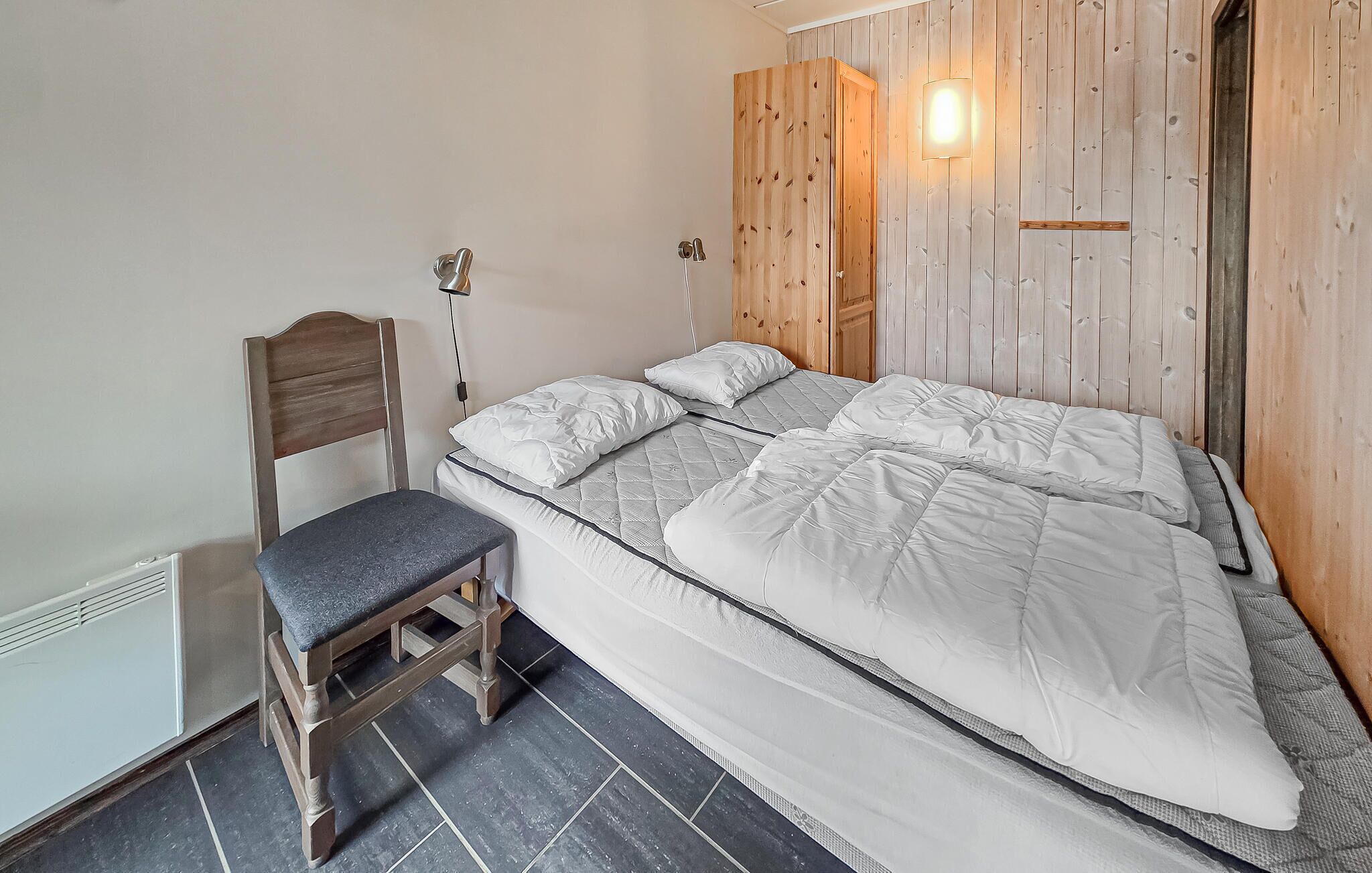 A single bed with a quilted mattress, a wooden chair with a cushion, a wall-mounted light, and a radiator.