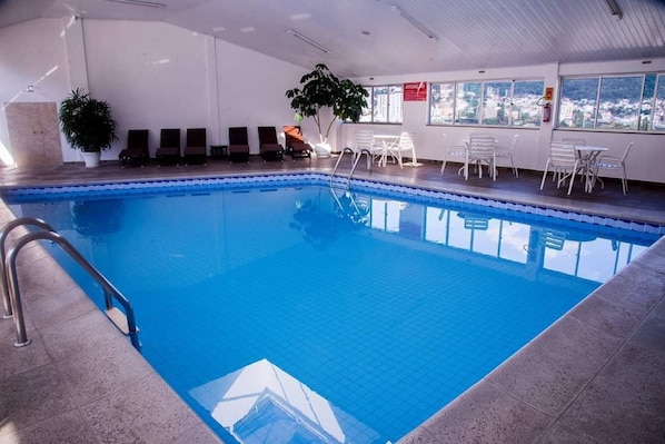 An indoor swimming pool with clear blue water, surrounded by lounge chairs and tables, with a view of a cityscape through large windows.