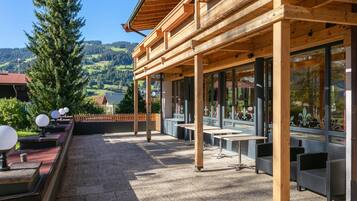 Ein Holzgebäude mit überdachter Terrasse, Sitzgelegenheiten im Freien und Blick auf eine gebirgige Landschaft.