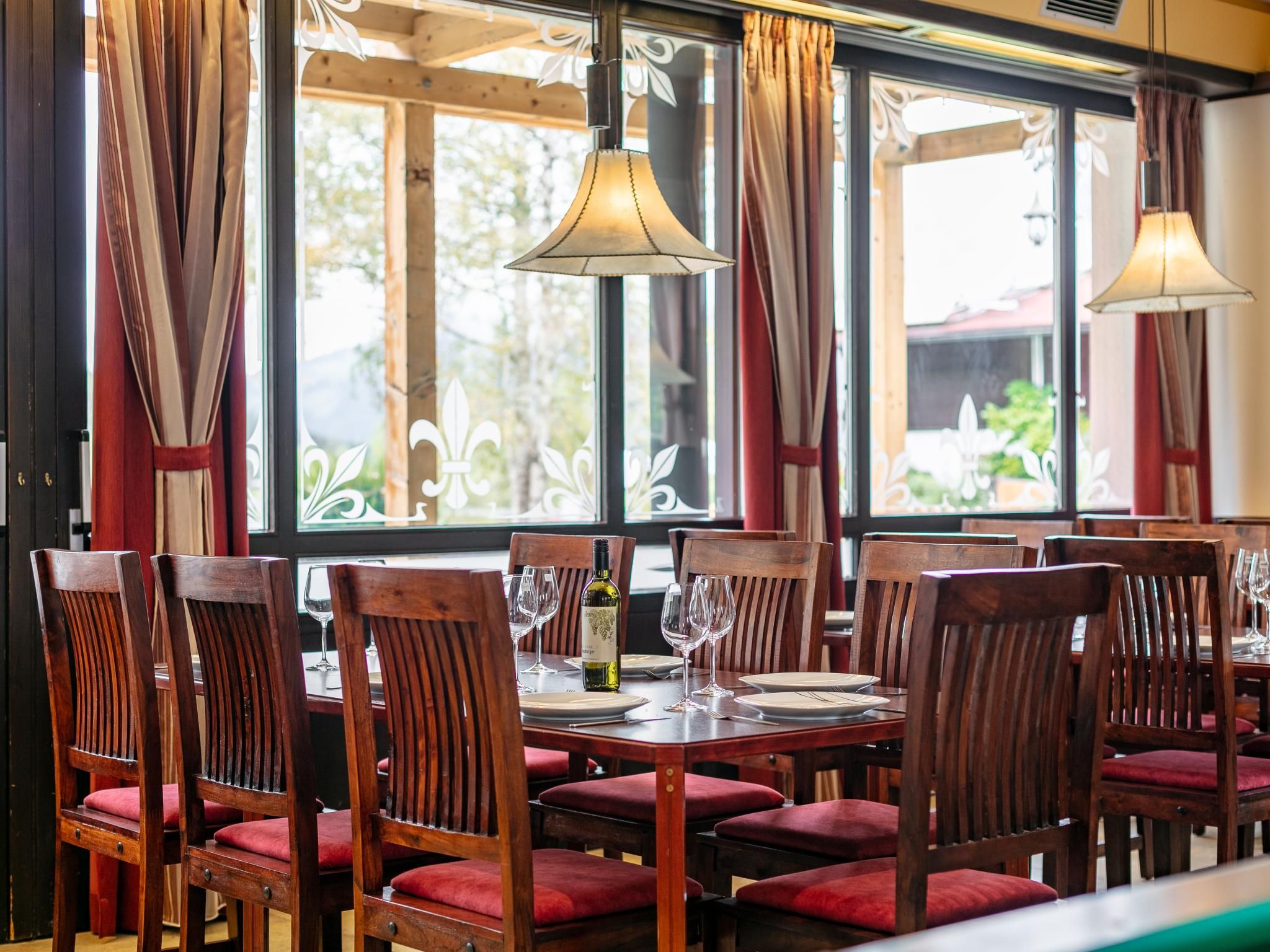 Ein Restaurantinneres mit Holztischen und -stühlen, großen Fenstern mit Vorhängen und hängenden Lampen.