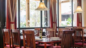 Ein Restaurantinneres mit Holztischen und -stühlen, großen Fenstern mit Vorhängen und hängenden Lampen.