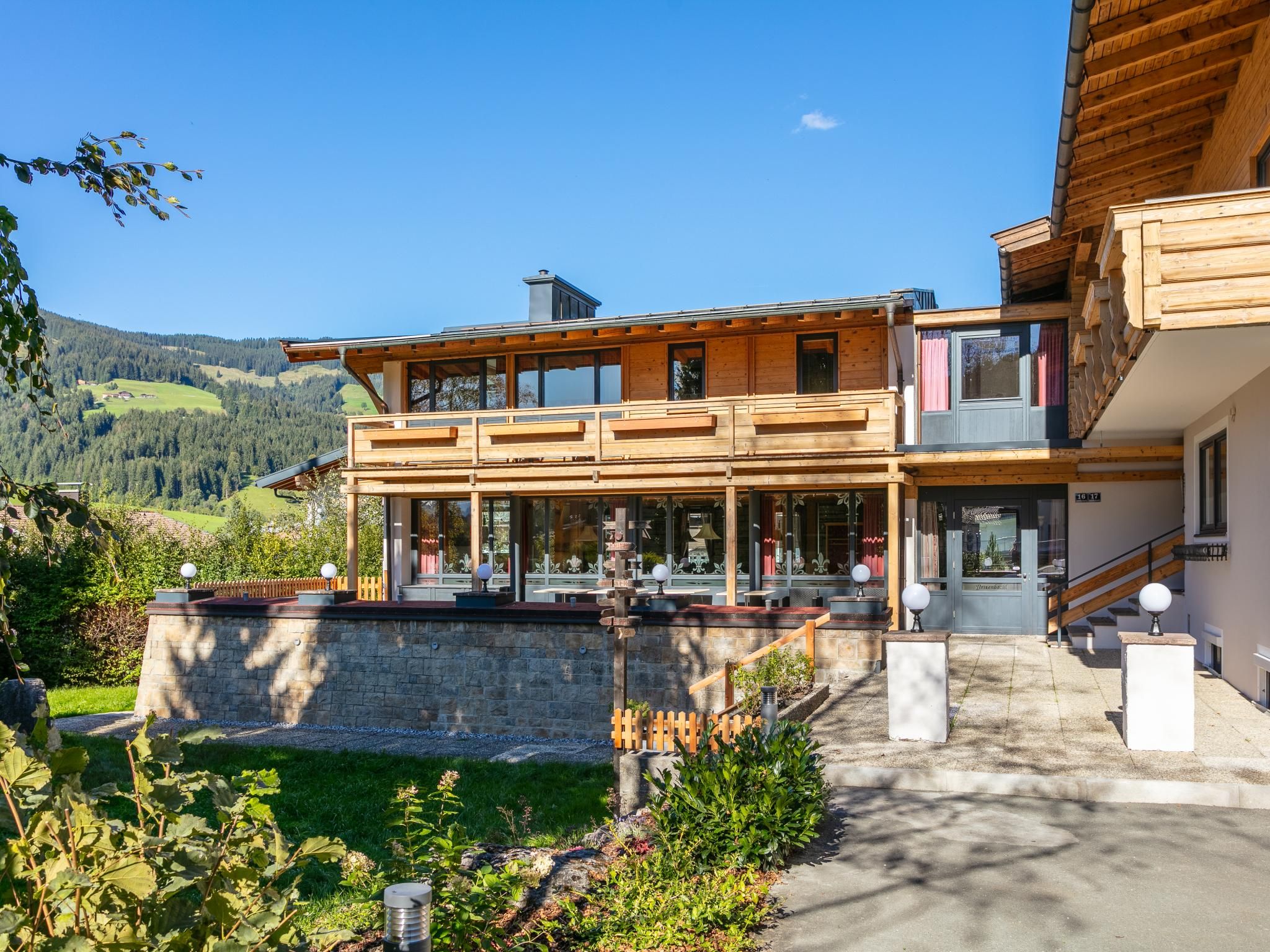 Une maison en bois à deux étages, avec un mur de pierre à l’avant, un balcon et une vue sur une vallée verdoyante.