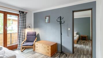 Une pièce comprenant un lit, une chaise, une commode, un porte-manteau et une vue sur l’extérieur, visible à travers une porte.
