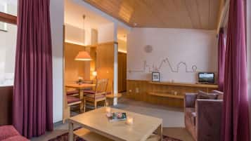 A modern hotel room with a wooden ceiling, a dining area with a table and chairs, a TV on a wooden shelf, and a cityscape wall art.