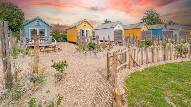 A row of colorful beach huts with a gravel path leading to them, surrounded by a wooden fence and greenery. - Fully equipped beach house with a terrace (Ellemeet)