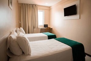 Ein Hotelzimmer mit zwei Betten, einem Schreibtisch mit Computer, einem an der Wand befestigten Fernseher und einem Fenster mit Vorhängen.