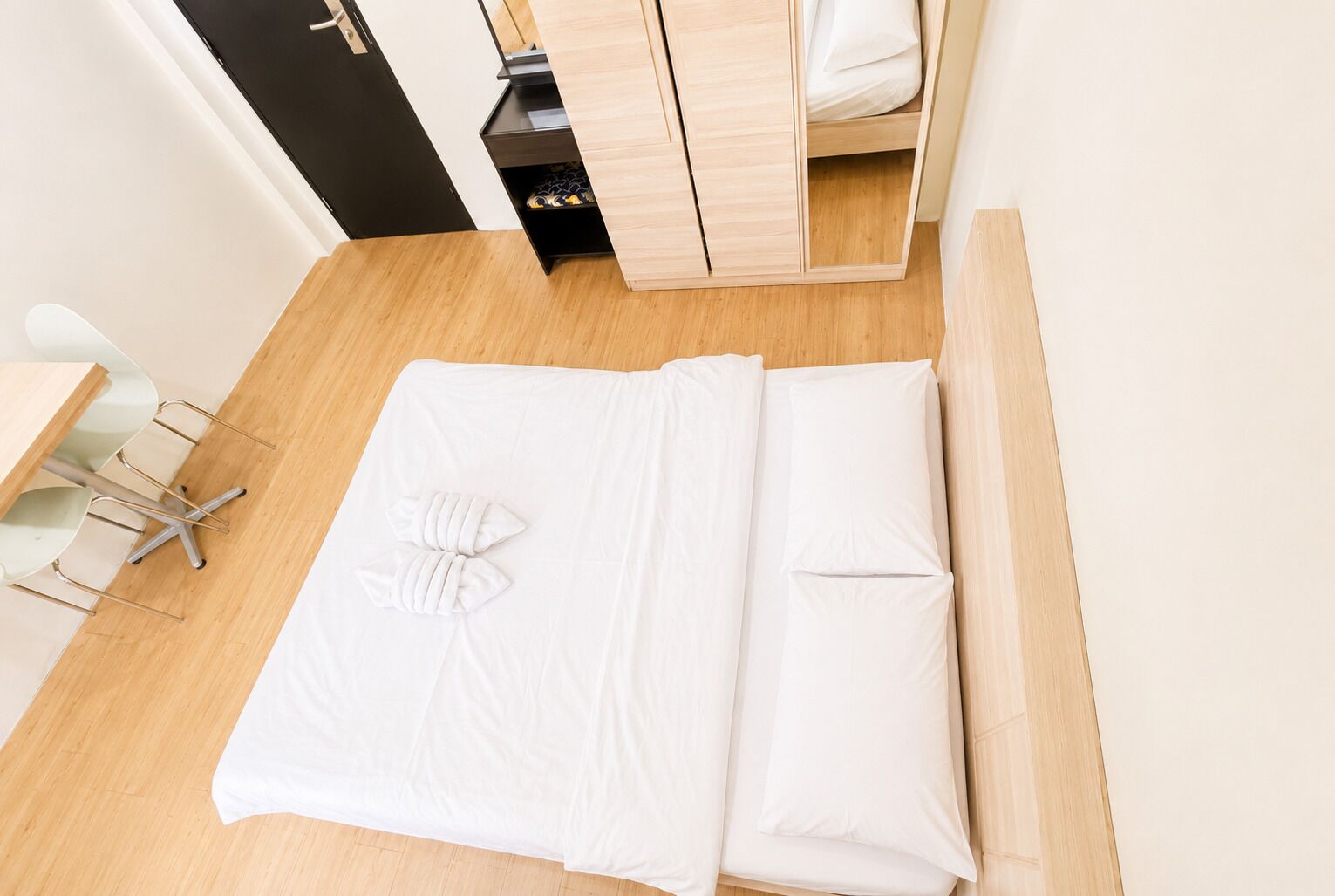 A neatly made bed with white linens and a decorative pillow, a wooden desk with a chair, and a small storage unit with shoes.