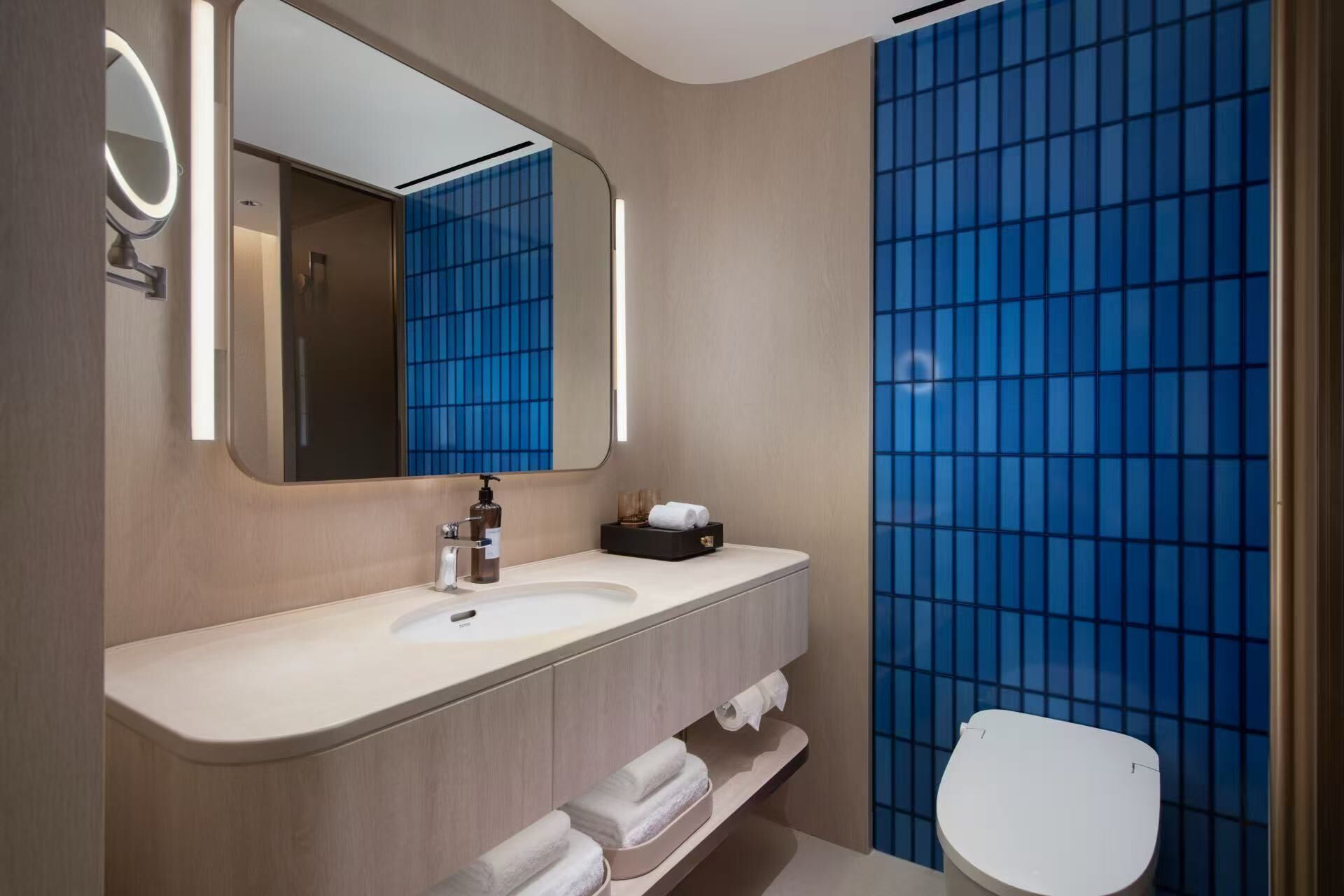 A modern bathroom with a large mirror, a sink, a toilet, and a blue-tiled shower wall.