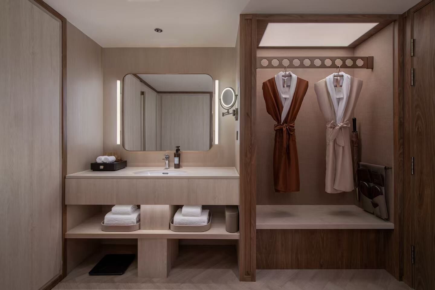 Een moderne badkamer met een houten wastafelmeubel, een grote spiegel en twee jassen aan een rek.
